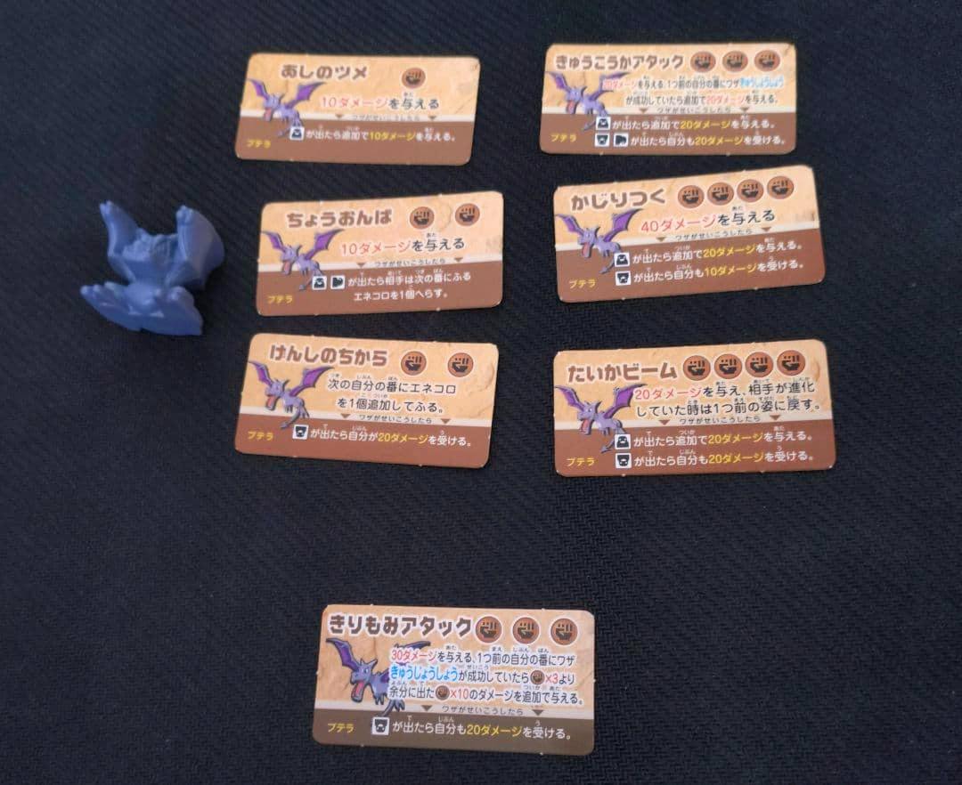 Blue plastic Ptera (プテラ) figure with several rectangular game cards arranged on a dark textured background.