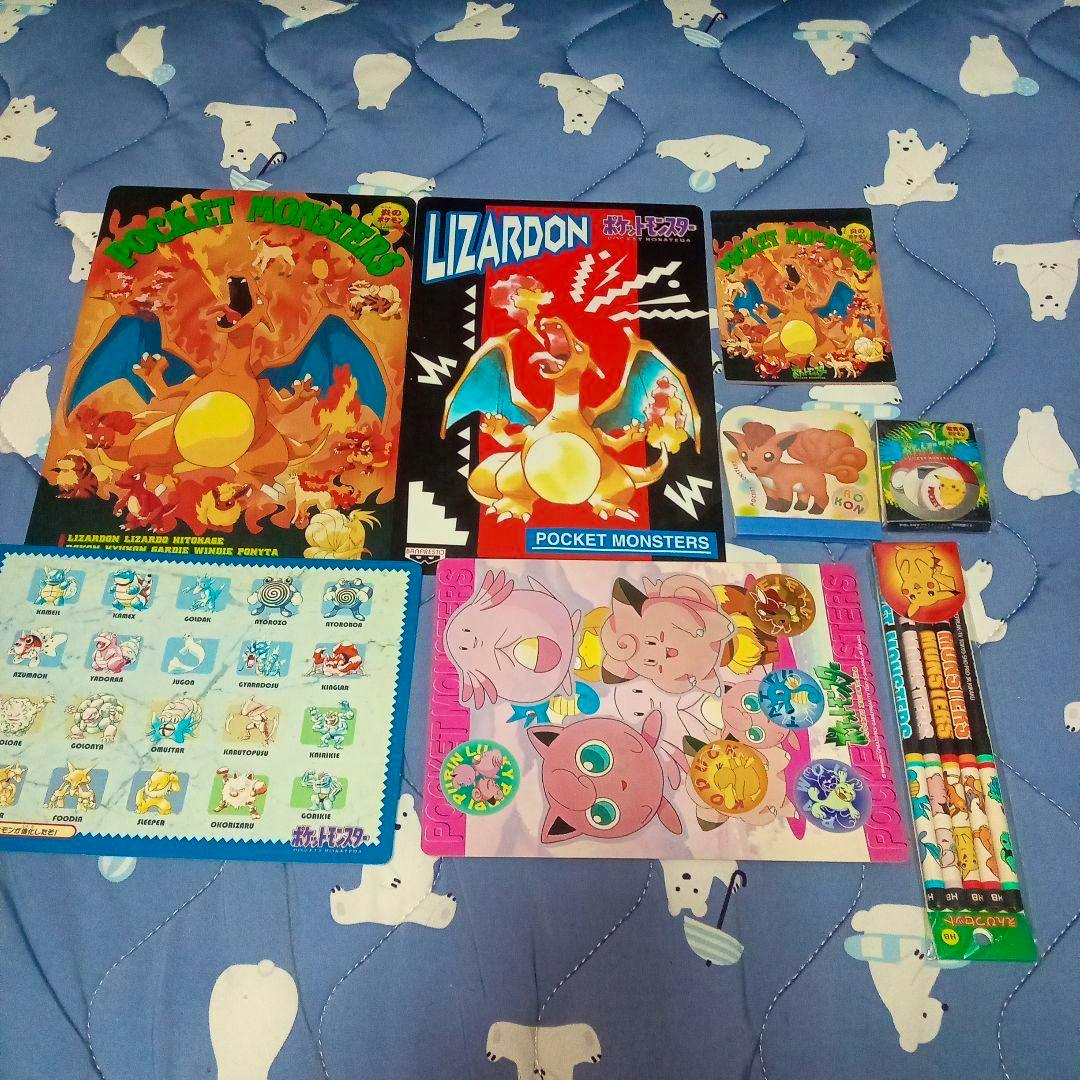 A collection of vintage Pocket Monsters stationery items on a blue quilted background, including Charizard-themed plastic mats, a Jigglypuff sheet, a sticker sheet, pencils, and small cards/erasers.