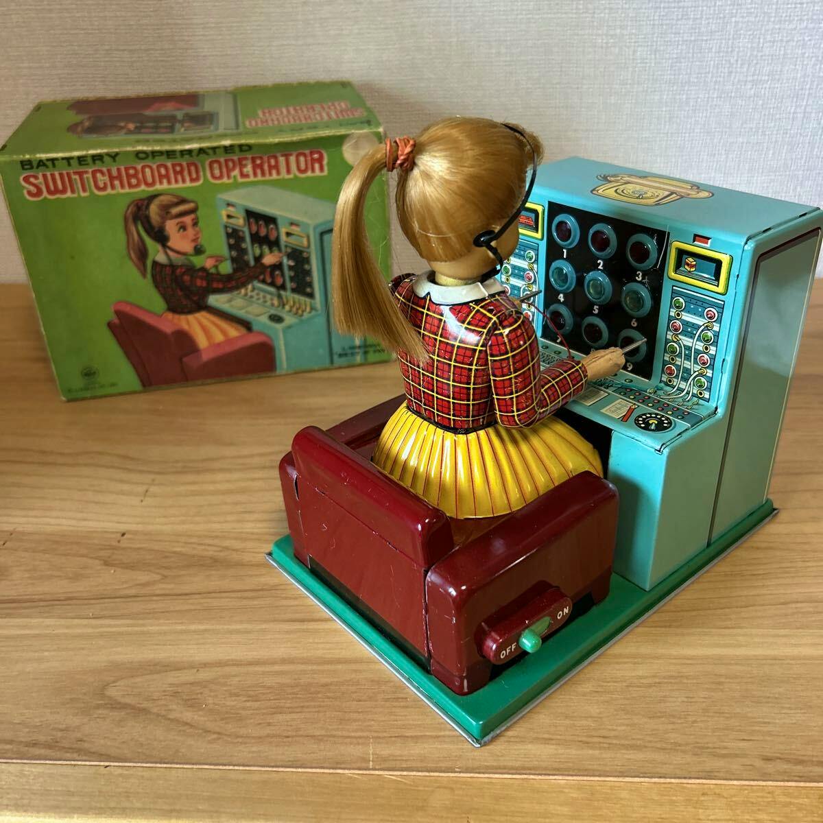 Tin switchboard operator toy seated at a teal switchboard with its original green box visible behind.