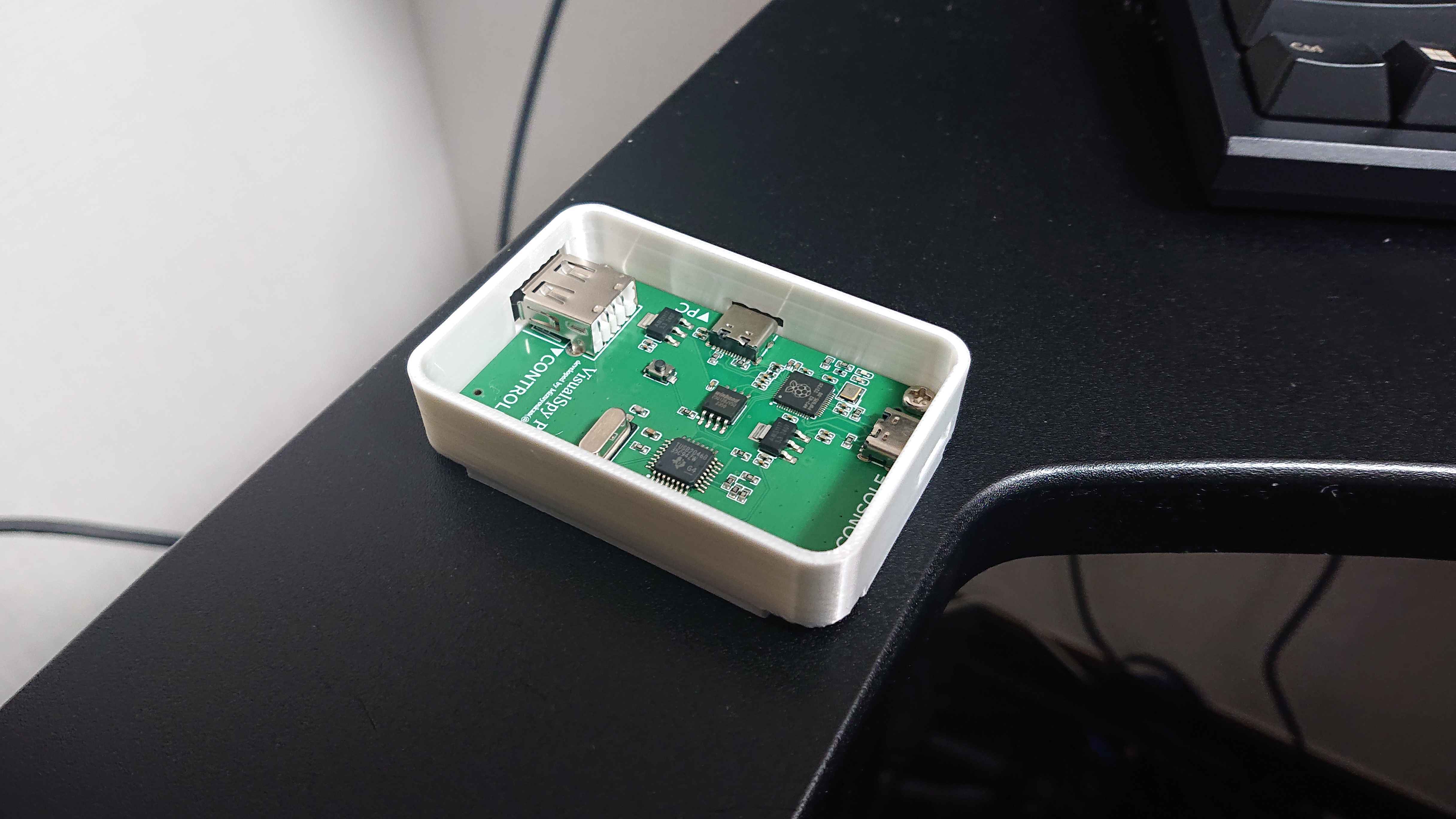A VisualSpy USB controller input visualizer board showing its green PCB and USB ports inside a white plastic enclosure on a desk.