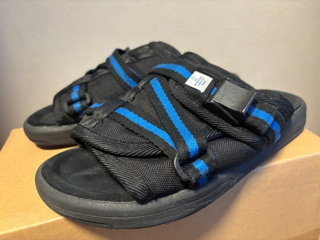 A pair of visvim CHRISTO sandals in black with blue stripes shown on a cardboard box from a front-side angle.