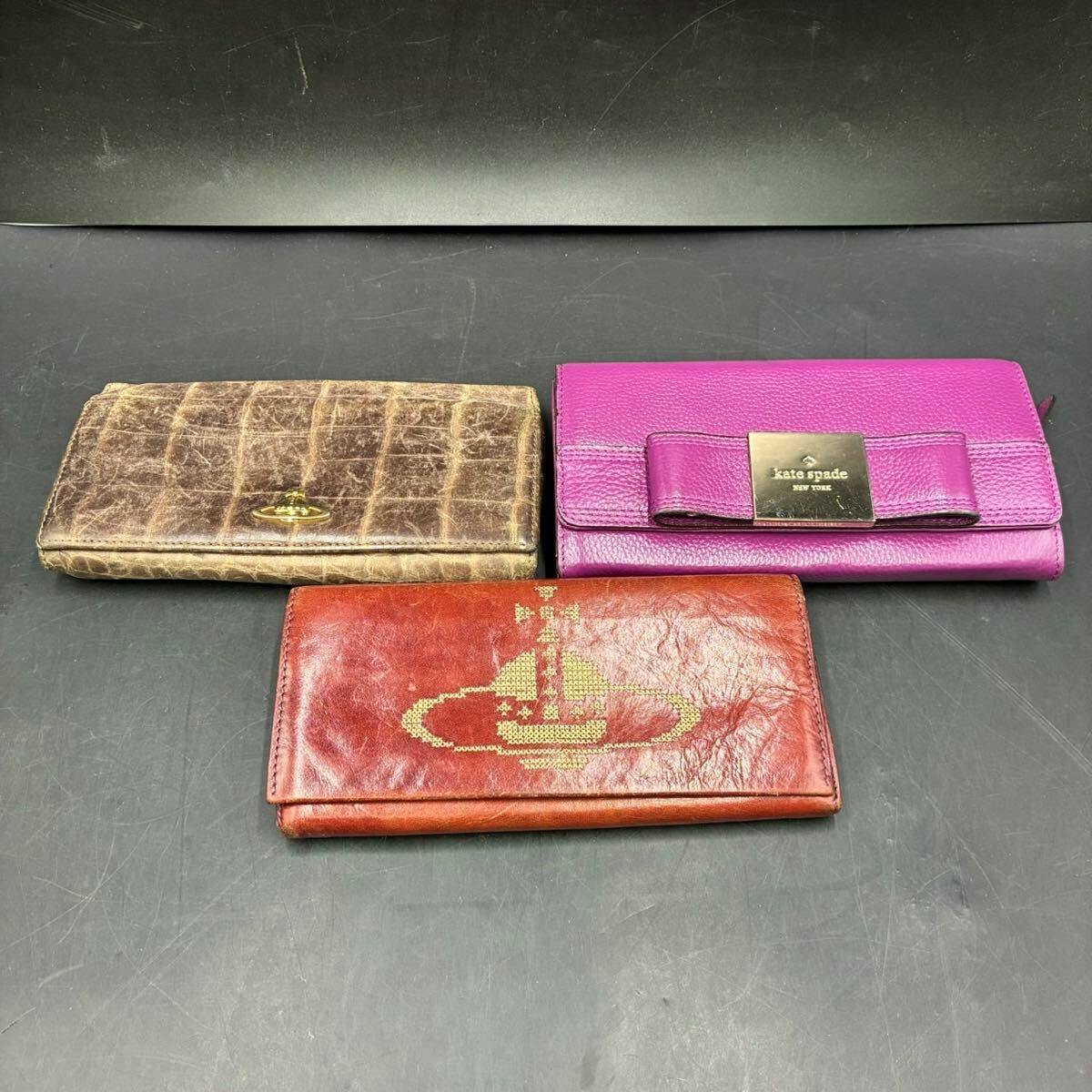 Three long leather wallets—brown croc-textured, purple Kate Spade, and red Vivienne Westwood—arranged on a dark surface.