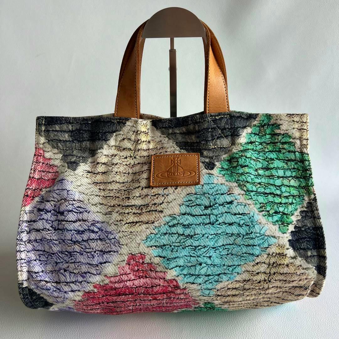 Front view of a multicolor canvas tote bag with tan leather handles and a leather orb logo patch.