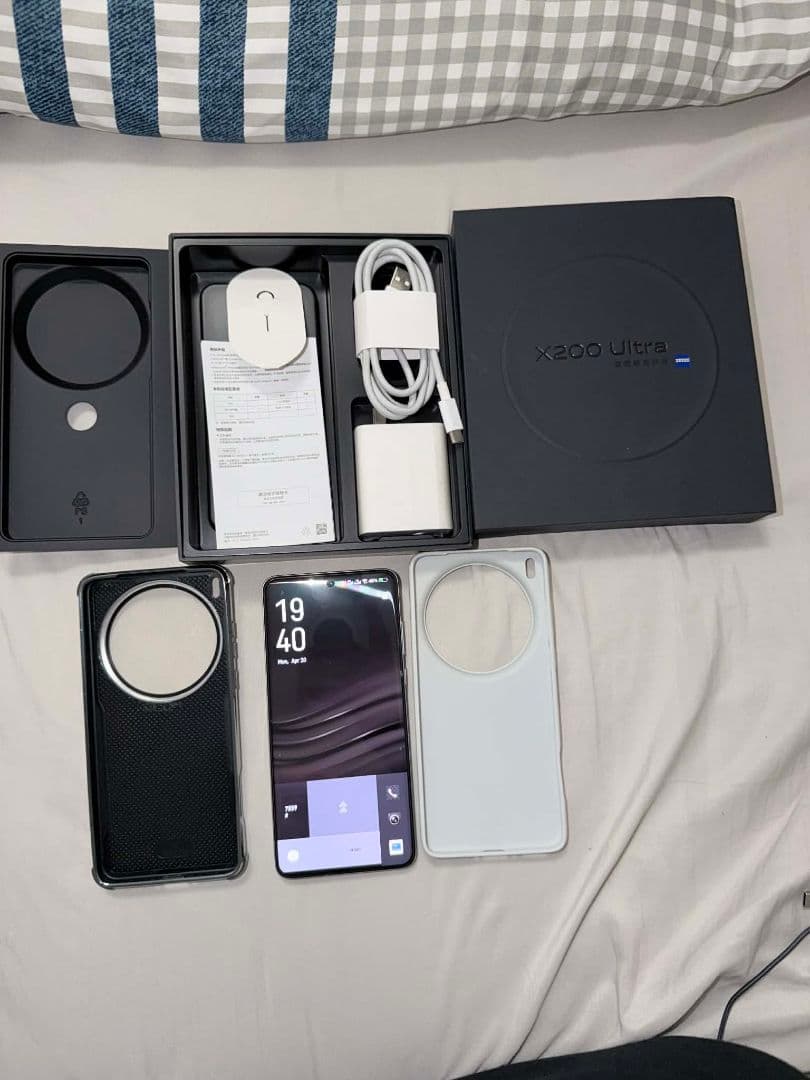 Vivo X200 Ultra smartphone shown with its box, charger, cable, paperwork, and two protective cases on a bed.
