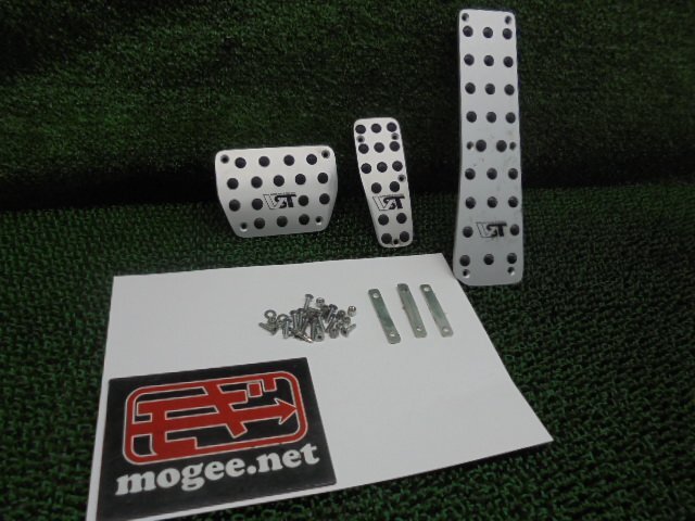Four silver car pedal covers with black raised dots and accompanying mounting screws and brackets displayed on a white sheet over a green textured background.