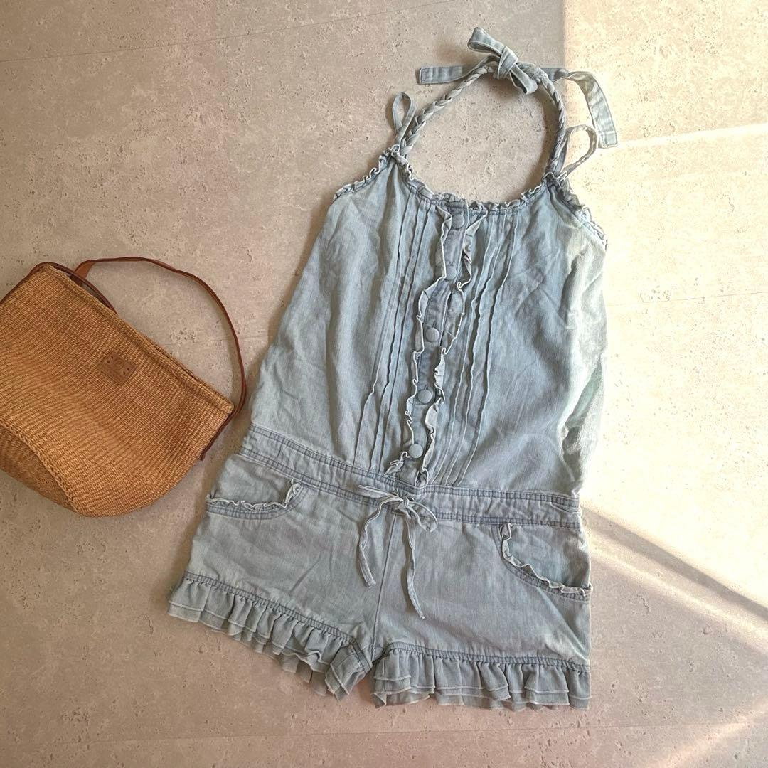 Light blue denim frilled short salopette (women's romper) laid flat next to a woven bag.