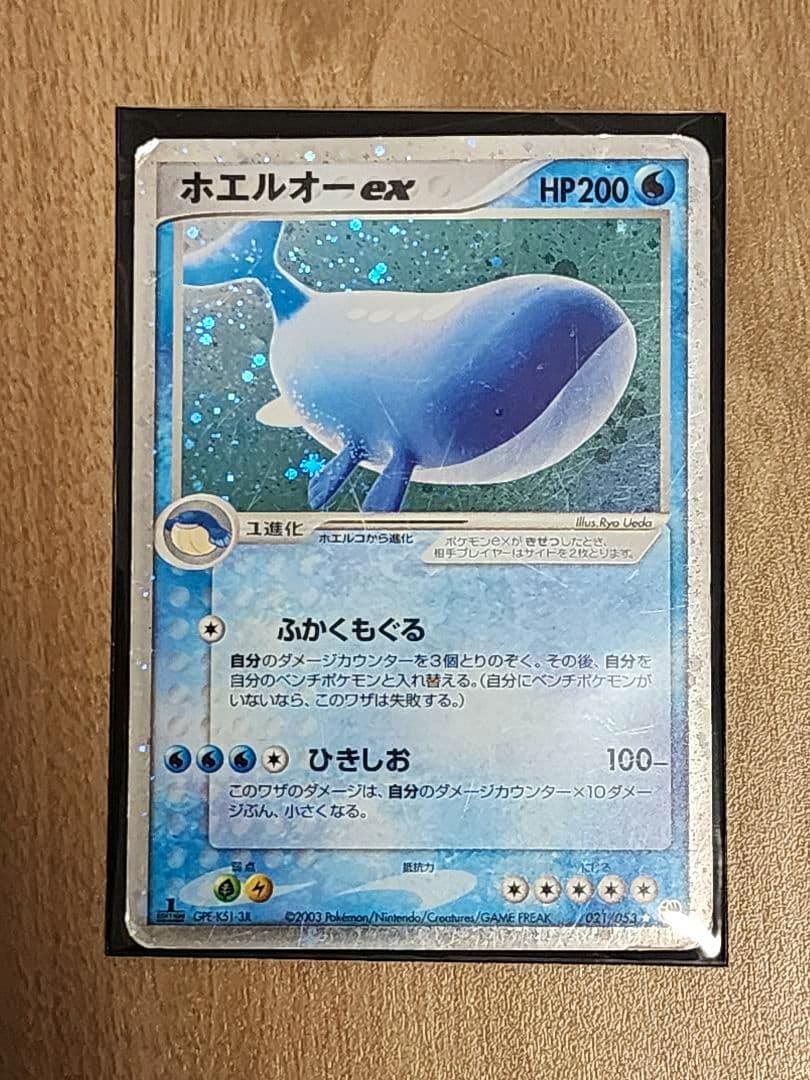 Front of the ホエルオー-ex Japanese trading card in a protective sleeve on a wooden surface, showing the card artwork and Japanese text.