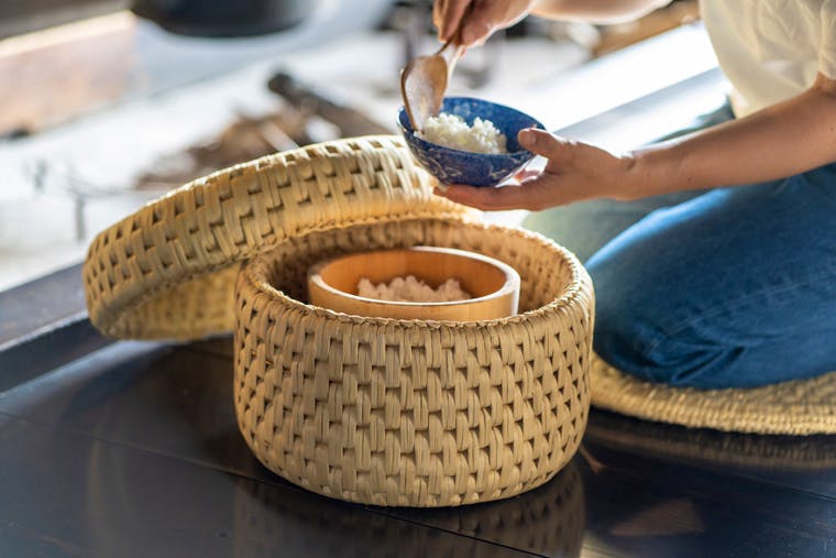 Woven straw basket with lid shown open holding a rice container while a person serves rice beside it.