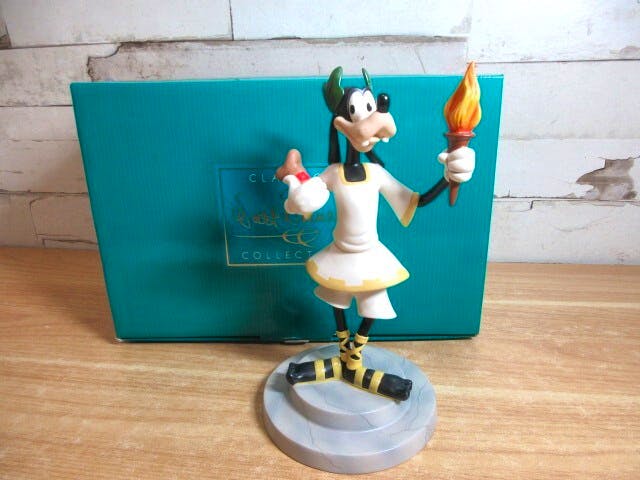 A WDCC Goofy Olympic Champ ceramic figure holding an Olympic torch on a round base with its box behind it.