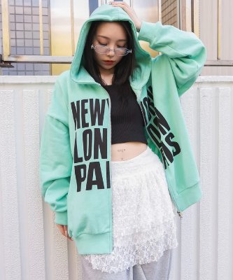 A mint-green zip-up hoodie with large black logo text worn by a model standing outdoors.