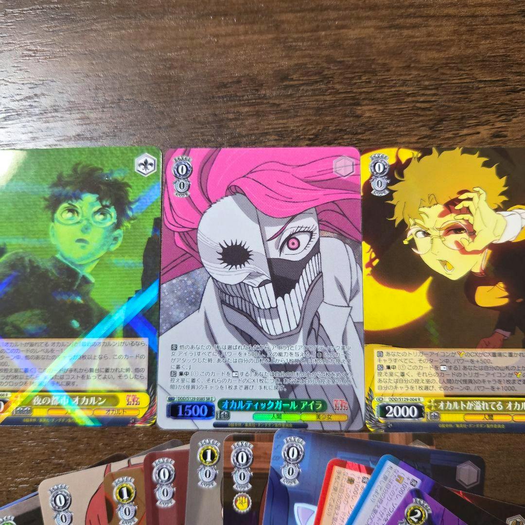Close-up of three Weiss Schwarz "Dandadan" trading cards laid side by side on a wooden surface with additional cards partially visible at the bottom.
