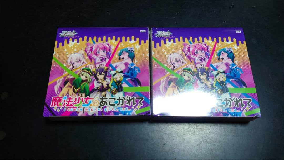 Two sealed booster boxes of ヴァイスシュヴァルツ 魔法少女にあこがれて shown side by side on a dark surface.