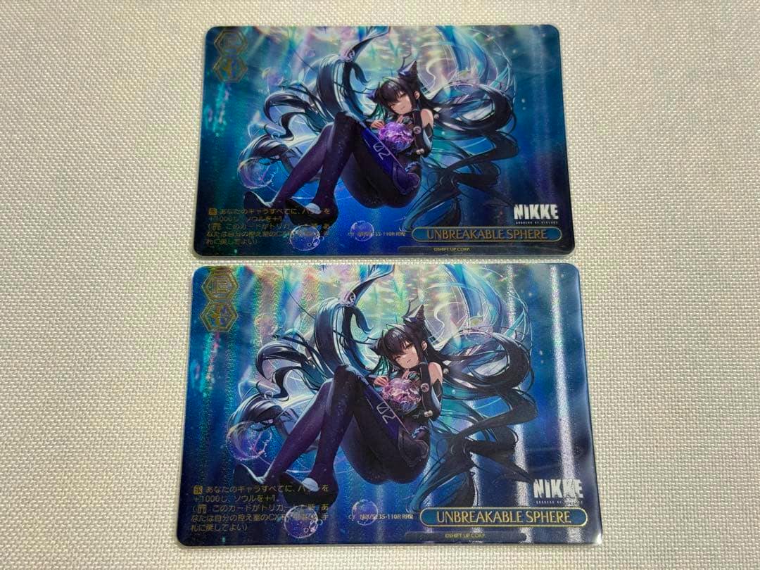 Photograph of two trading cards face-up showing the NIKKE "UNBREAKABLE SPHERE" illustrated artwork and card title.