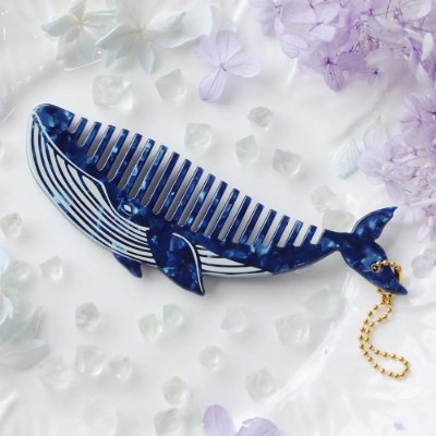 A blue-and-white whale-shaped comb with a small gold chain charm, shown on a white surface with crystals and purple flowers.