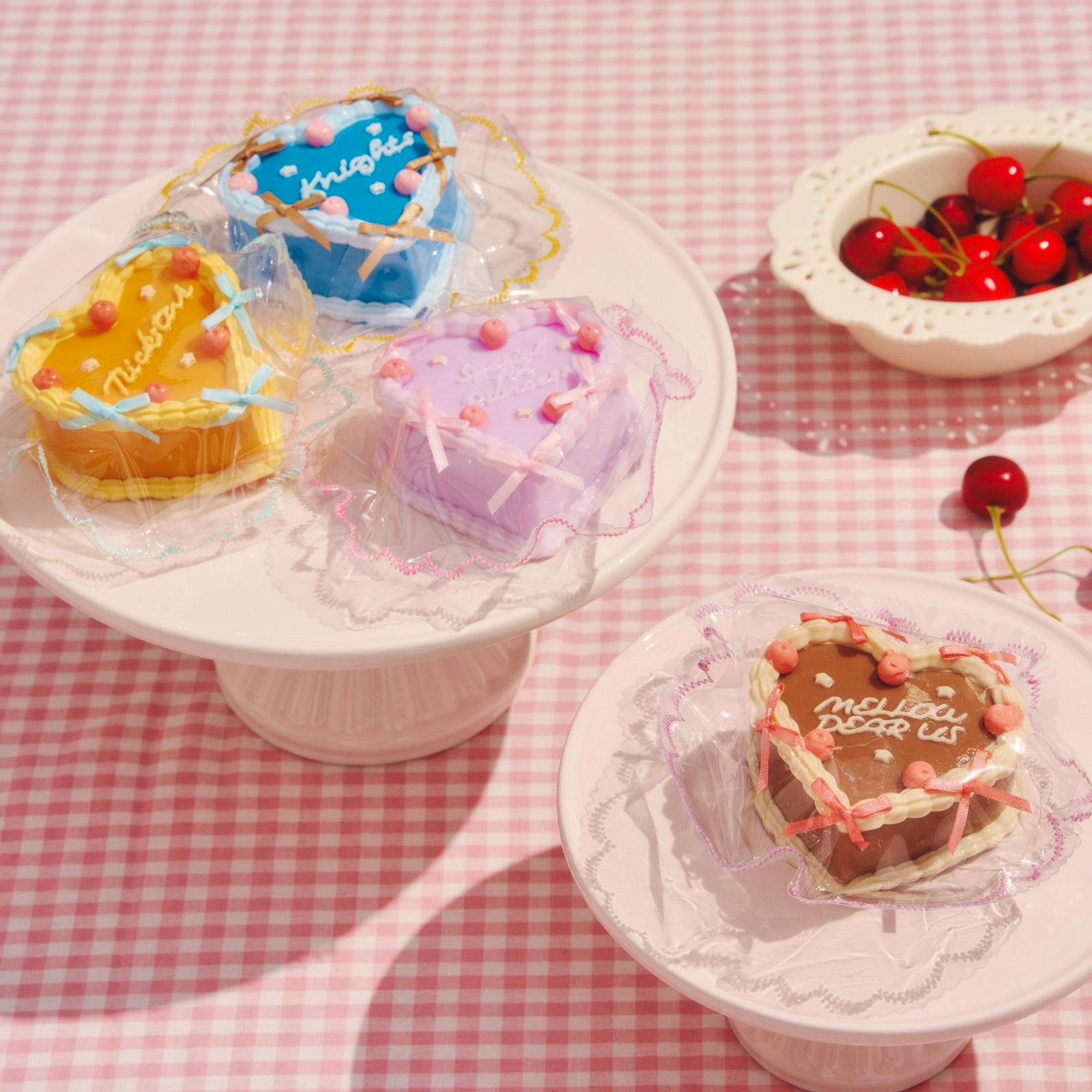 Three pastel heart-shaped cake squishy toys wrapped in clear plastic on white cake stands over a pink gingham background.