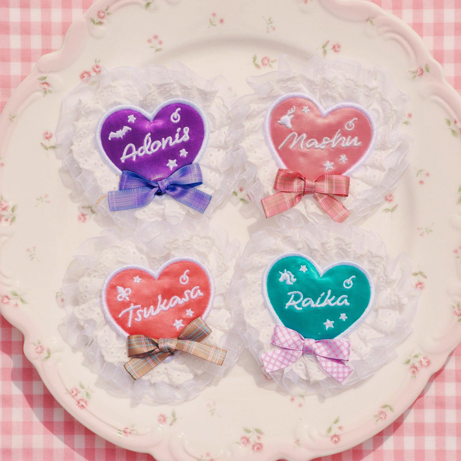 Photo of four heart-shaped idol name badges with lace frills and ribbon bows arranged on a decorative plate.