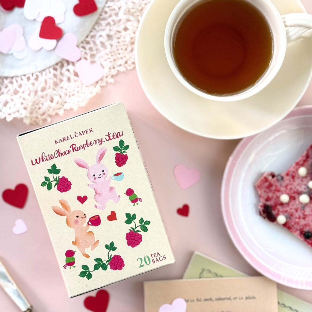 A box of White Chocolate Raspberry tea bags sits beside a cup of brewed tea and a plate with a dessert on a pink background.