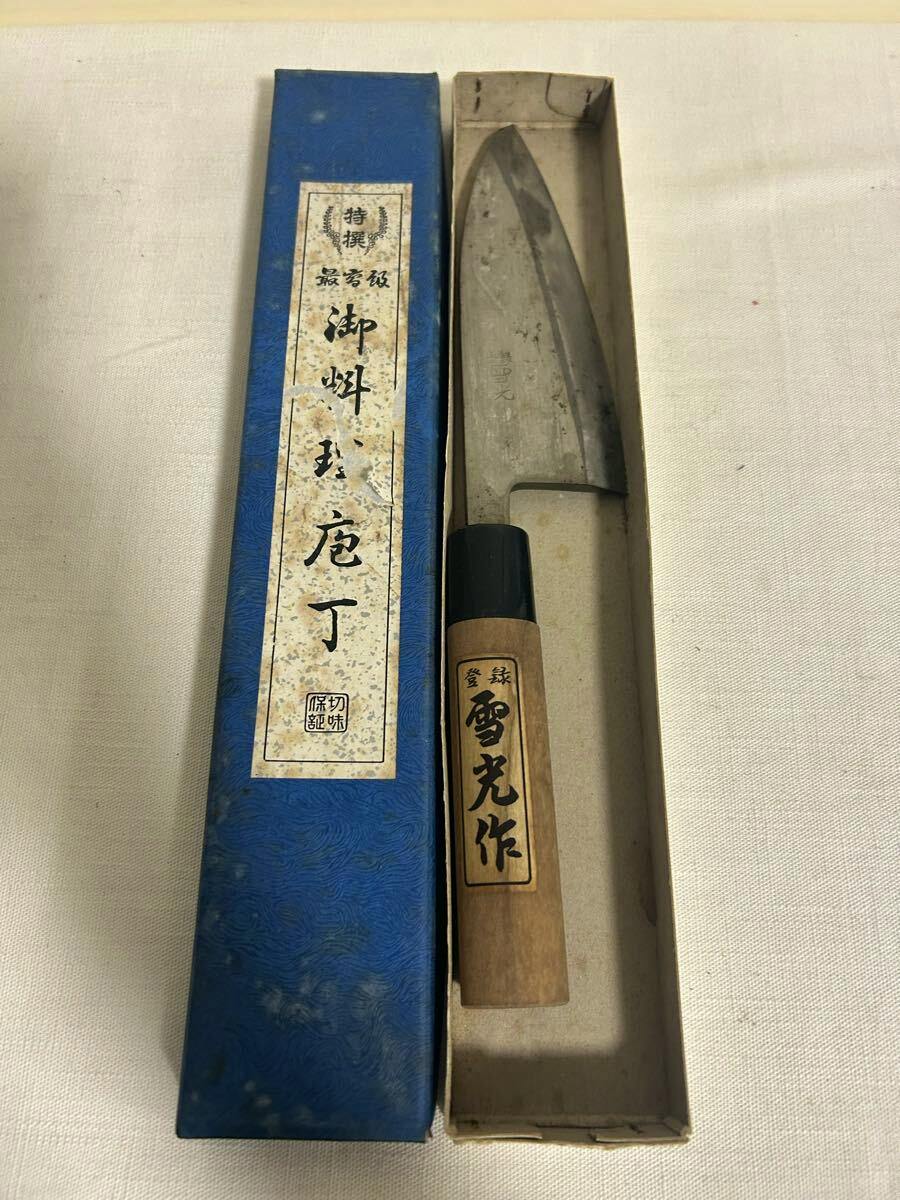 A chef knife with a wooden handle and metal blade displayed inside its box next to a blue lid with a Japanese label.
