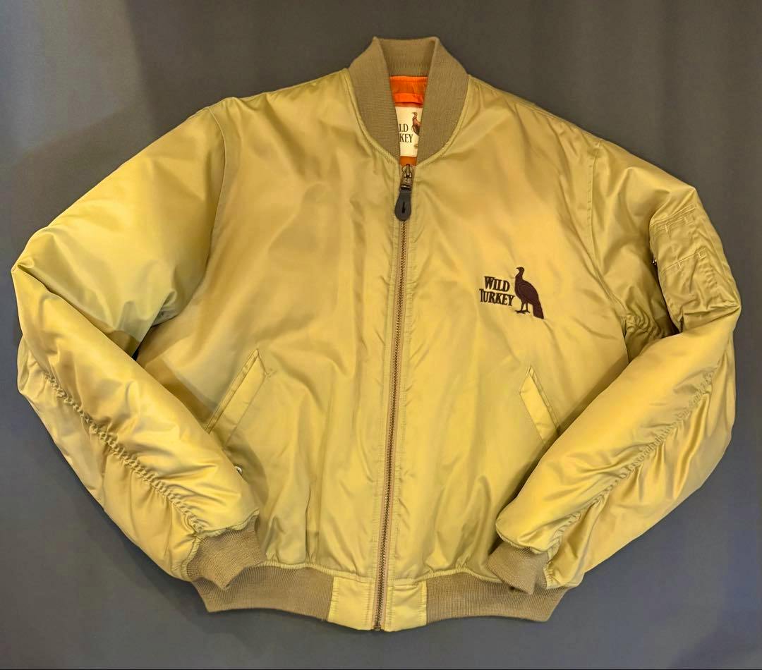 Front view of the WILD TURKEY MA-1 flight jacket in light tan polyester with a center zipper, ribbed collar and hem, and a small Wild Turkey logo with bird silhouette on the chest.