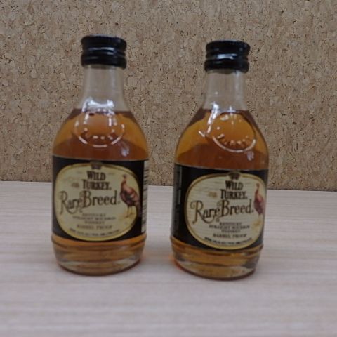 Two Wild Turkey Rare Breed 50ml mini whiskey bottles side by side on a table with a corkboard background.