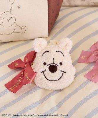 Front view of the Pooh face pouch charm (polyester) showing embroidered facial features on a small plush cream pouch, placed on a striped surface beside decorative ribbons.