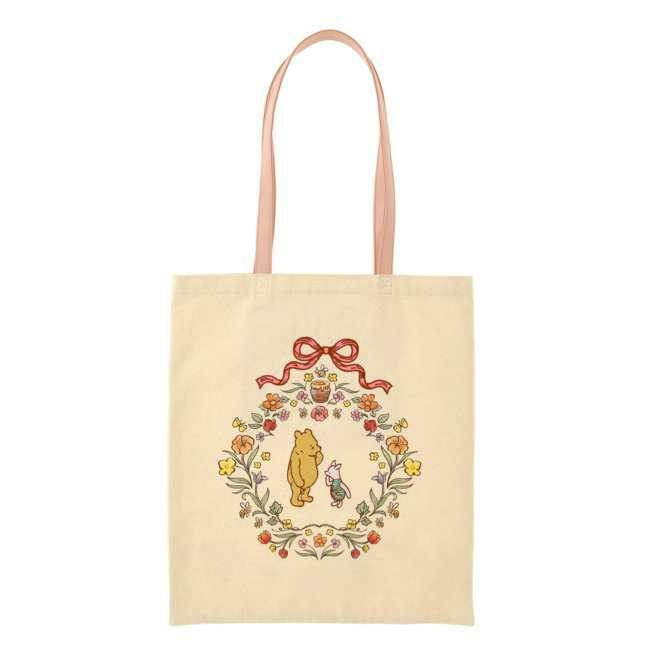 Beige cotton tote bag with pink handles featuring Pooh and Piglet inside a printed floral wreath on the front.