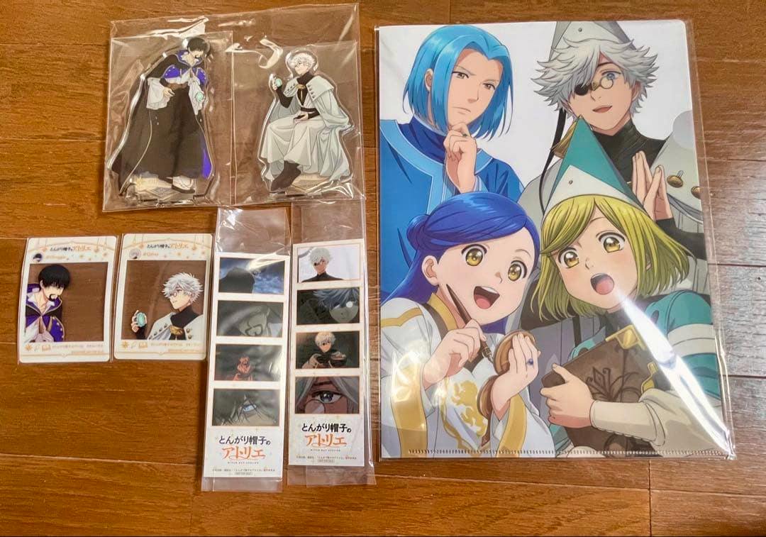 Photo of multiple とんがり帽子のアトリエ goods on a wooden floor, including two acrylic character stands, small character cards, film-strip style bookmarks, and a large illustrated clear file.