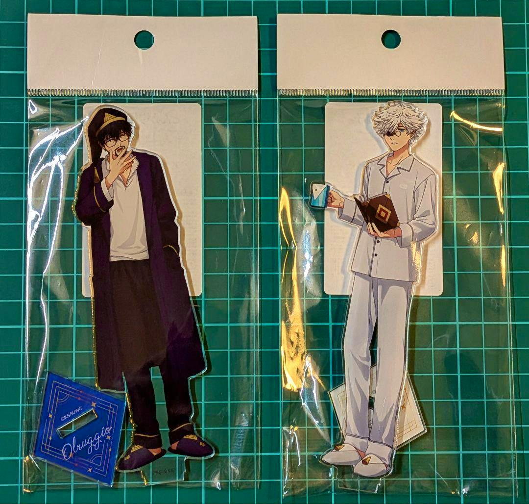 Two packaged acrylic character stands from とんがり帽子のアトリエ shown side-by-side on a green grid cutting mat.