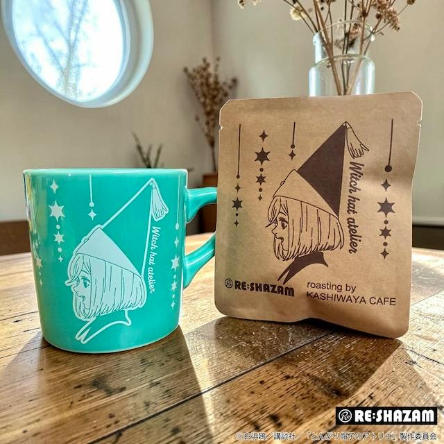 Photograph of a turquoise ceramic mug printed with a Witch Hat Atelier illustration next to a packaged drip coffee sachet on a wooden table.