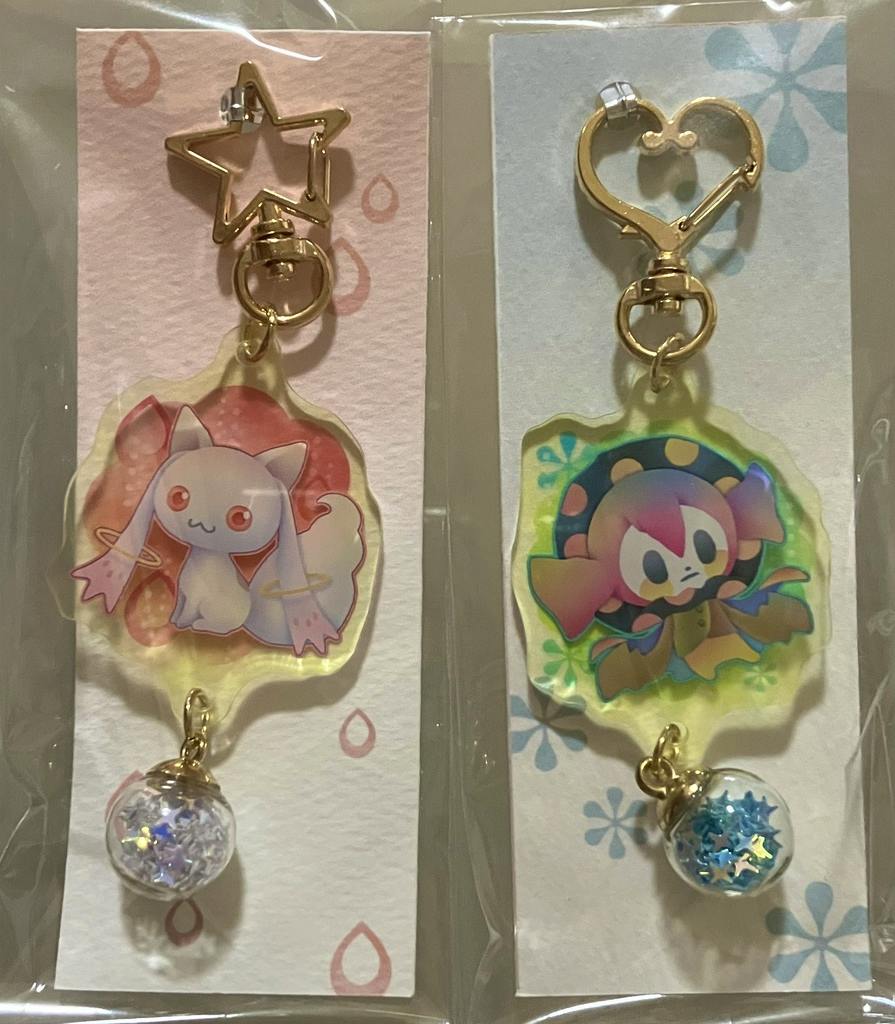 Two acrylic keychains shown in their packaging, each with a character charm, a gold star or heart clasp, and a small clear globe filled with star-shaped glitter.