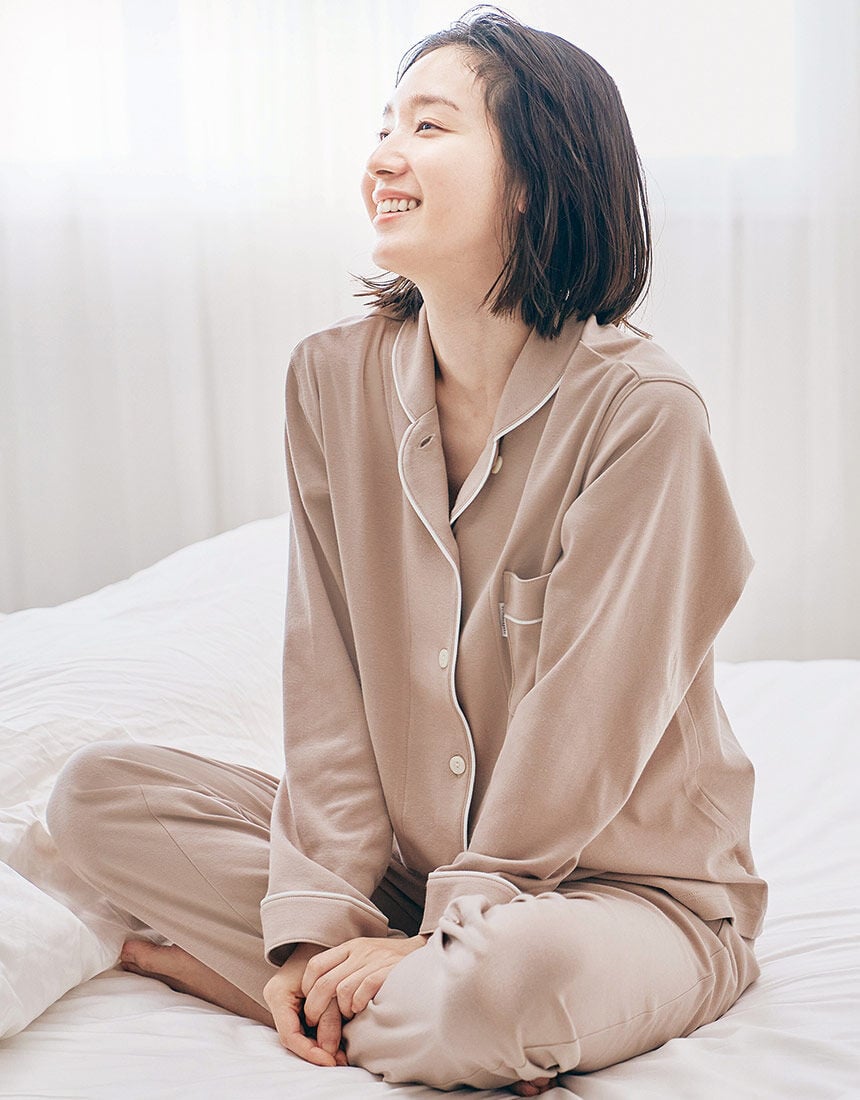 A woman sitting on a bed wearing beige women's cotton pyjamas with contrast piping.