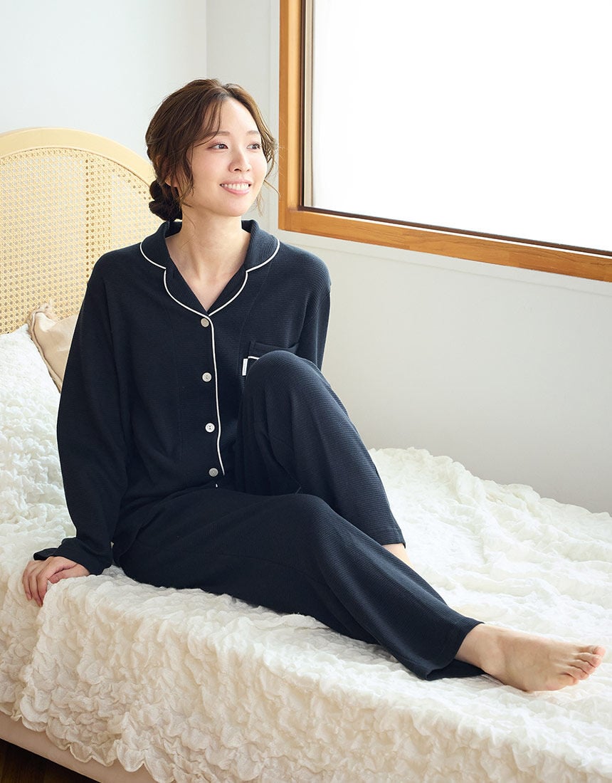 A woman sitting on a bed wearing navy long-sleeve, button-up women’s cotton pyjamas with matching pants.