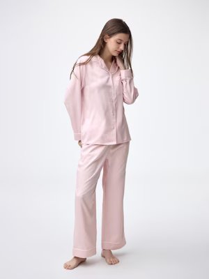 Woman wearing pink satin long-sleeve top and long pants pajama set on a white background.
