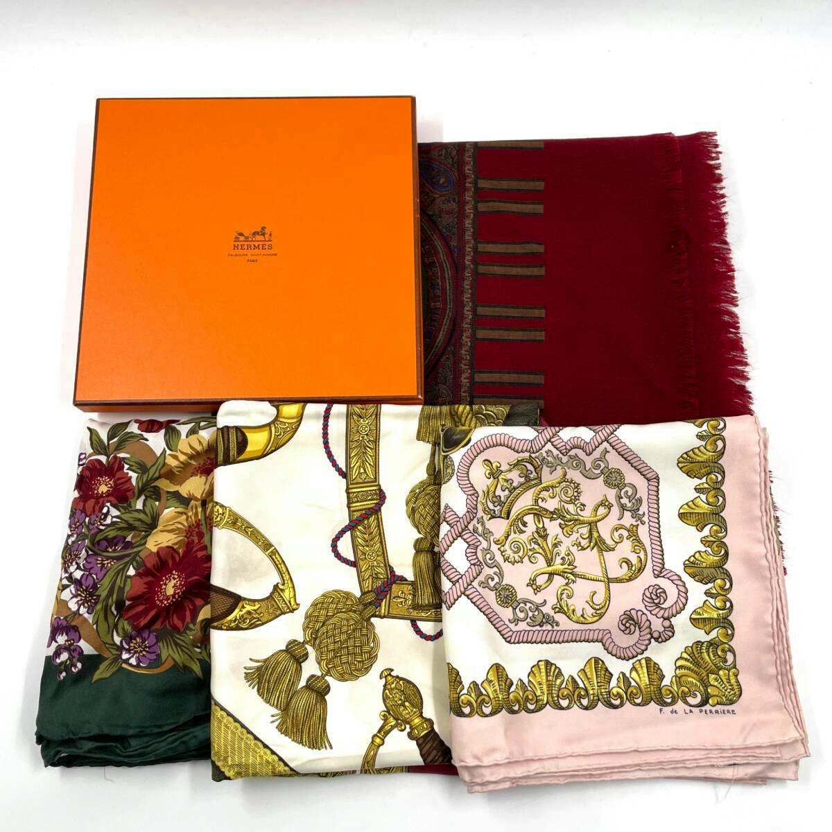 Four folded scarves (silk) including patterned pieces and a red scarf arranged with an orange Hermès gift box on a white background.