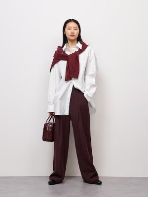 Woman modeling the wide tuck pants in dark red, styled with a white oversized shirt and a matching sweater draped over her shoulders.
