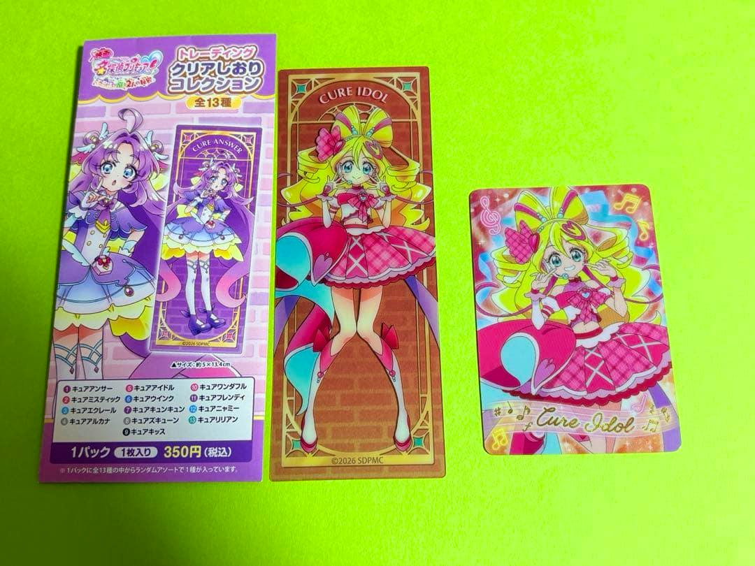 Photo of the product on a green surface showing printed packaging at left, a long rectangular clear bookmark featuring Cure Idol in the center, and a small glitter card with the same character at right.
