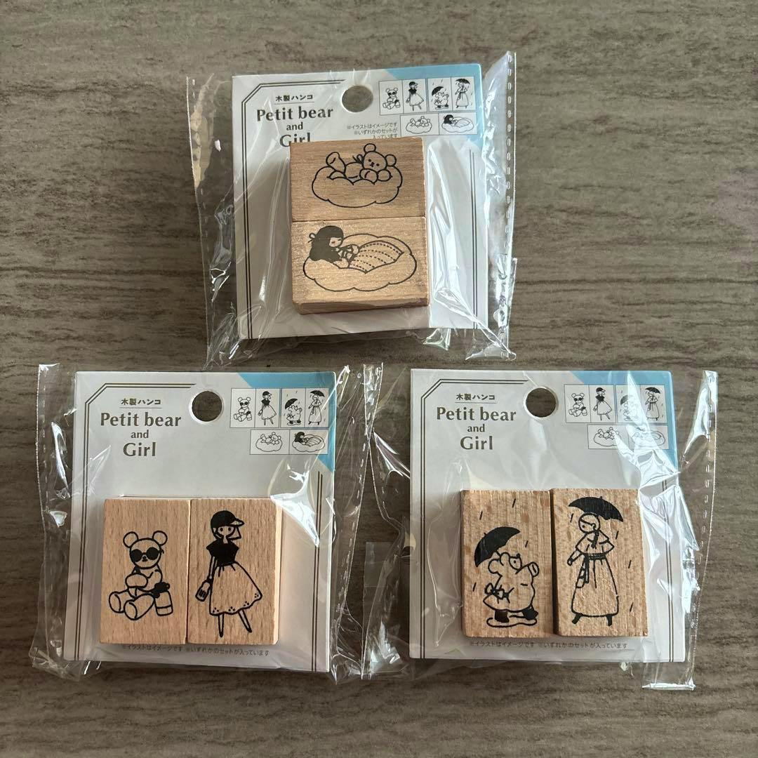Three packaged wooden stamps showing Petit bear and girl designs arranged on a wooden surface.