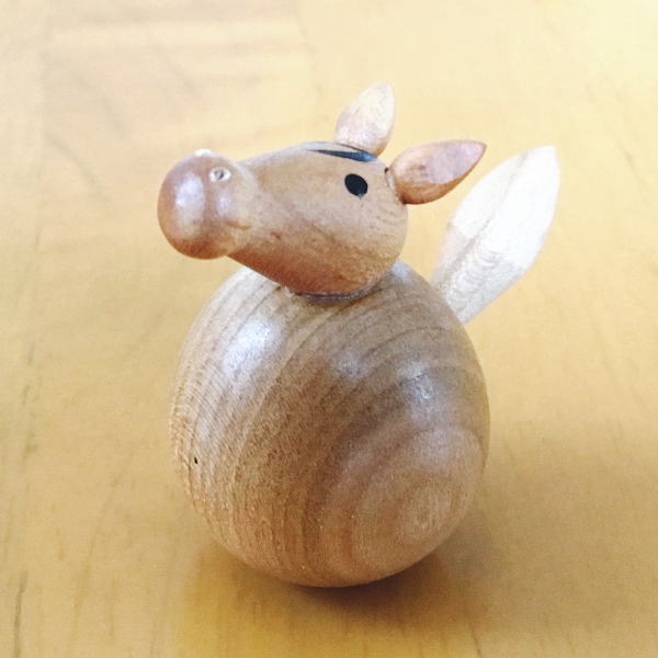 A small wooden horse-shaped roly-poly ornament with a rounded body, carved head, and a white tail resting on a light wooden surface.