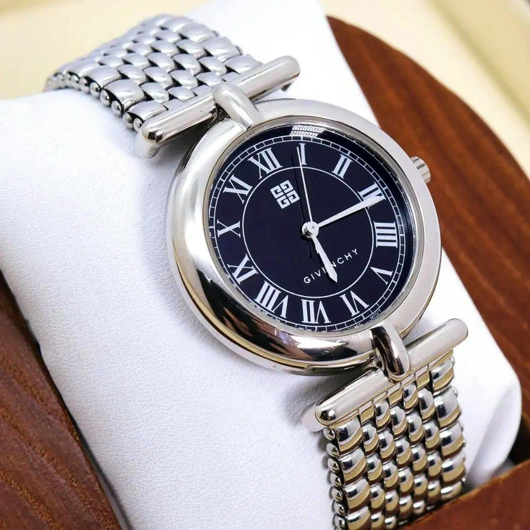 Close-up of a Givenchy round metal-case wristwatch with a black dial, Roman numerals, and a metal bracelet.