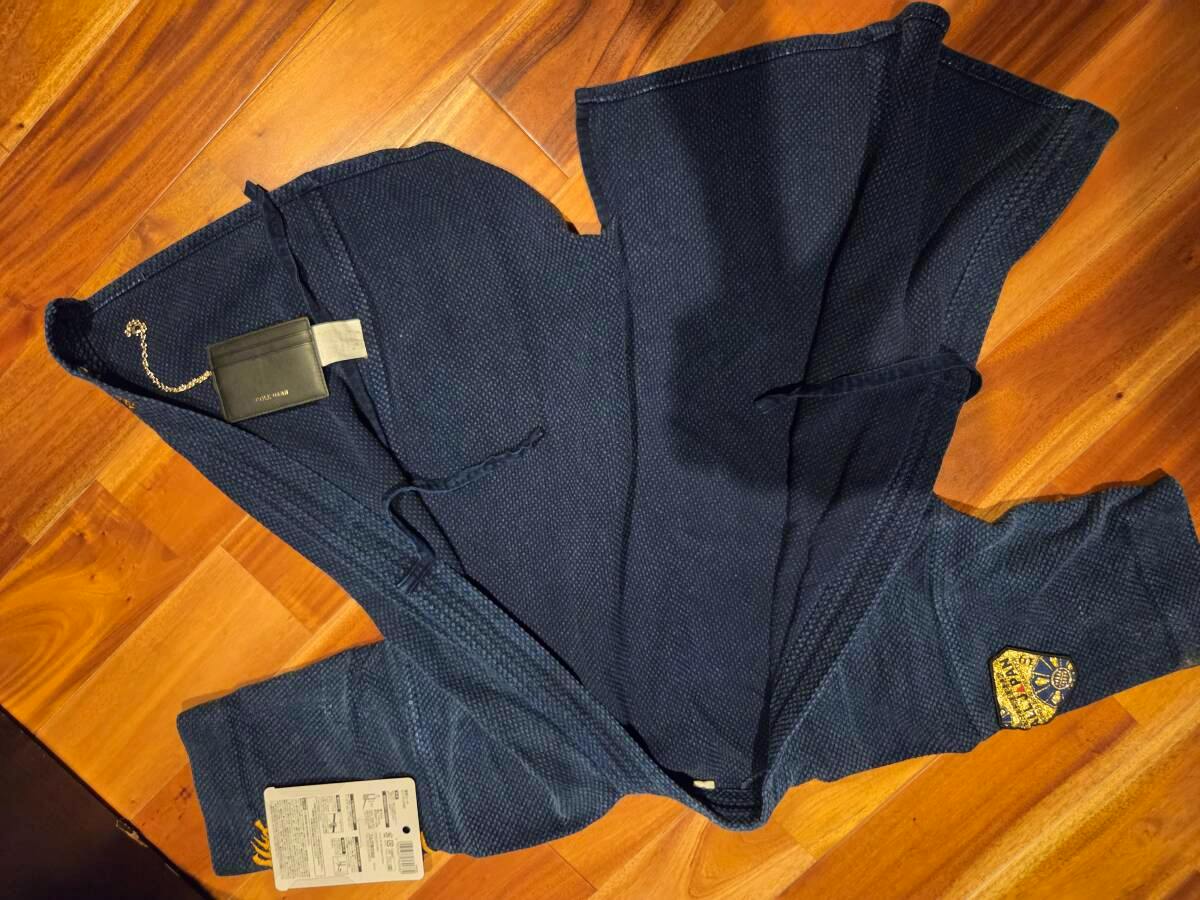 A navy indigo woven kendo jacket laid flat on a wood floor, showing the front, a sleeve patch, and attached tags.