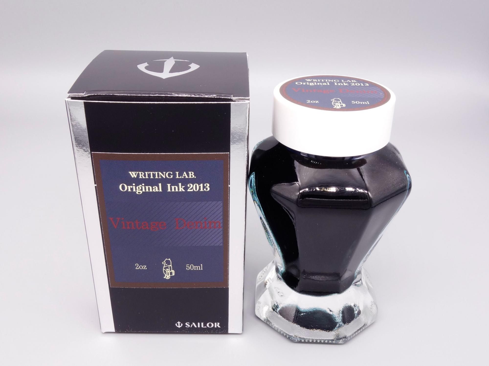 A glass bottle of WRITING LAB Original Ink (Vintage Denim) with a white cap displayed next to its box on a white background.
