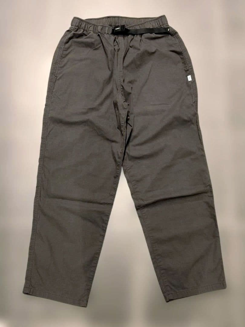 Flat-lay front view of the WTAPS men's trousers laid flat, showing an elastic waistband with buckle and a small side label.