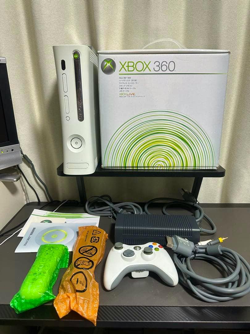 An Xbox 360 console standing next to its retail box with a controller, power brick, cables, and manuals laid out on a table.