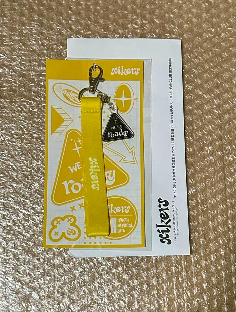 Yellow Xikers fanclub keychain with a yellow fabric strap and a small triangular black charm on its branded backing card.