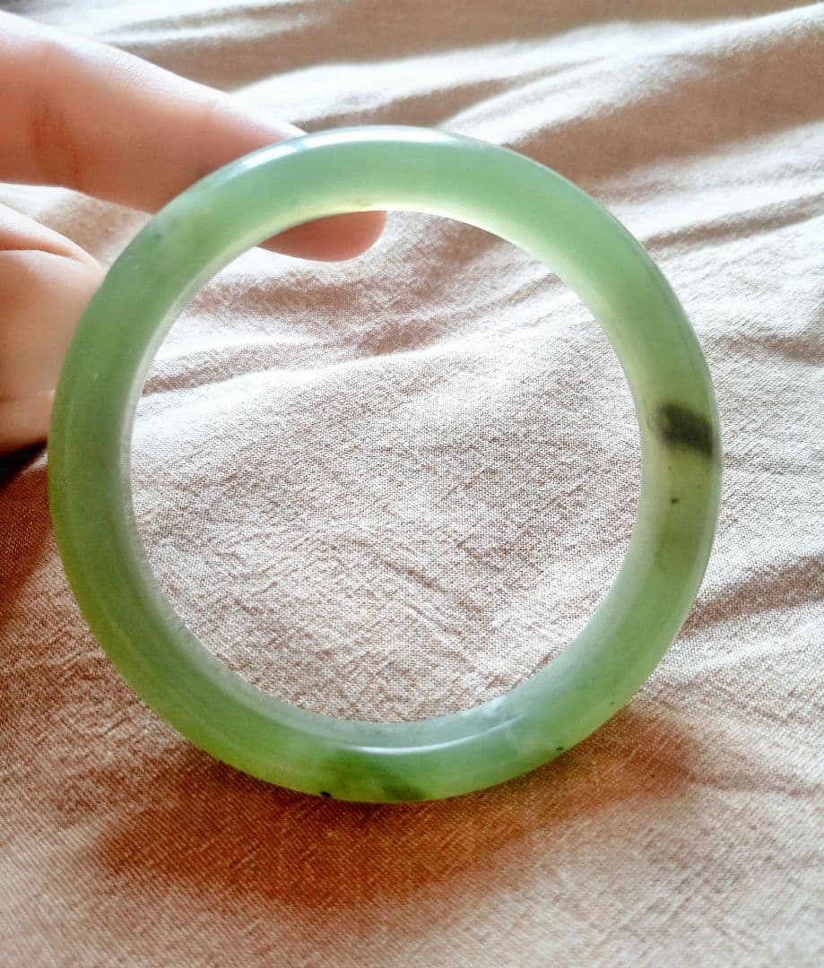 Green jade bangle bracelet held by a hand over a beige fabric background.