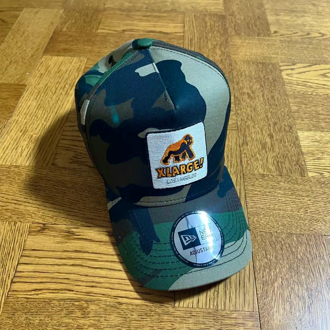 Camo XLARGE New Era snapback hat with an embroidered XLARGE patch and an adjustable New Era sticker on the brim, photographed on a wood floor.