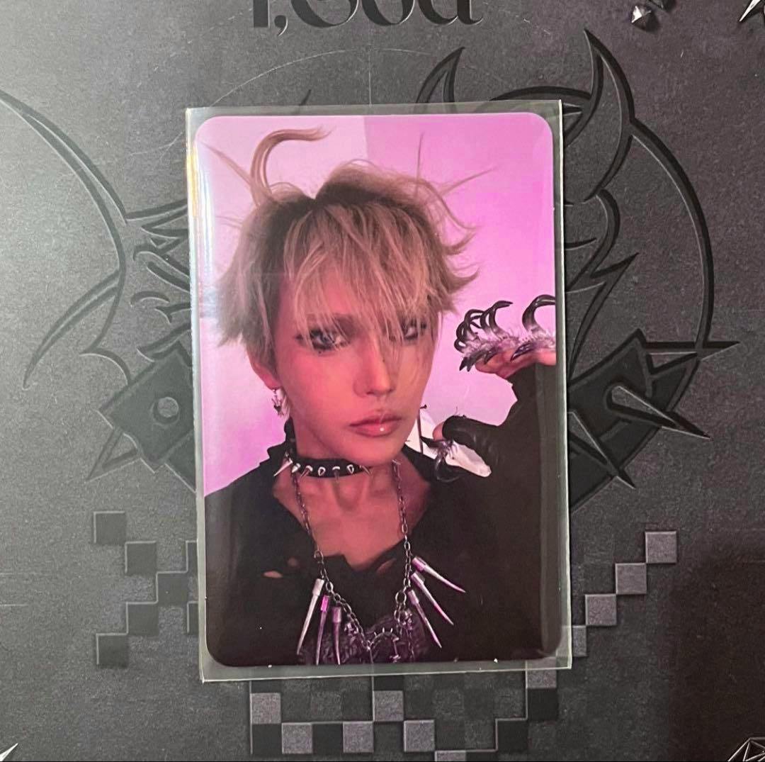 Photocard in a clear sleeve showing a blond-haired person wearing a choker and spiked necklace against a purple-pink background on a dark patterned surface.