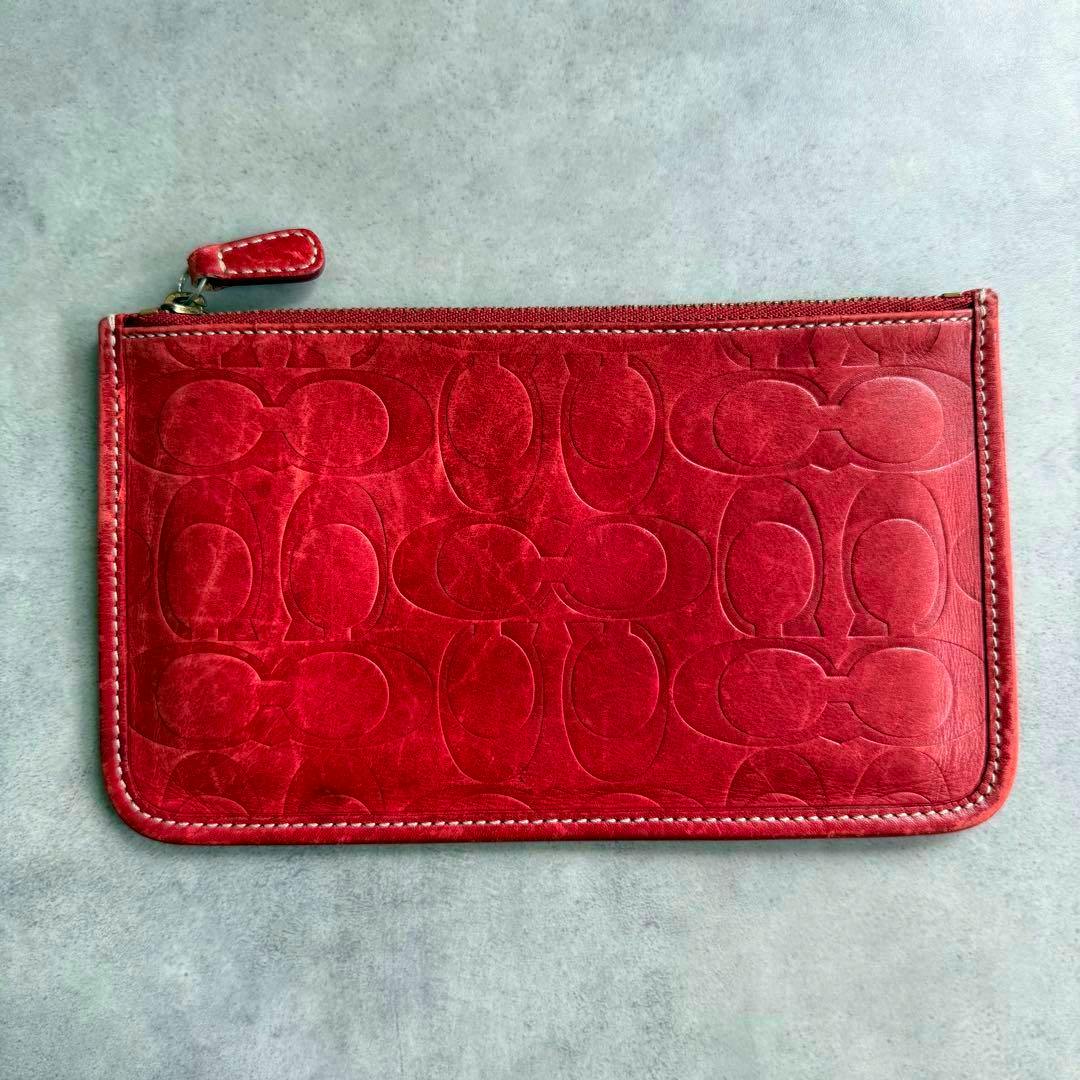 Red COACH embossed cow leather pouch with signature pattern and a top zipper pull laid flat on a gray surface.