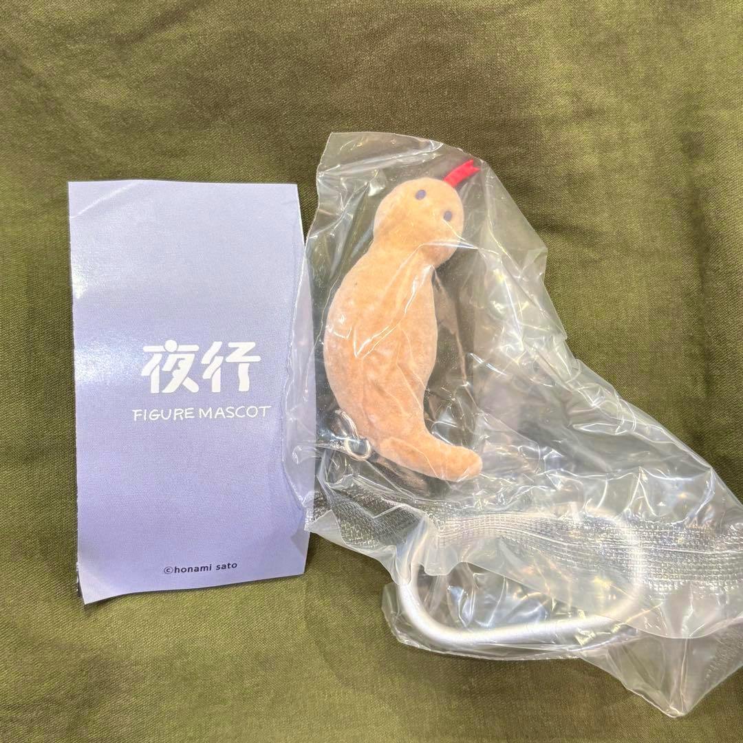 A small Tsuchinoko plush figure mascot sealed in clear plastic beside a purple "夜行 FIGURE MASCOT" insert on a green cloth background.
