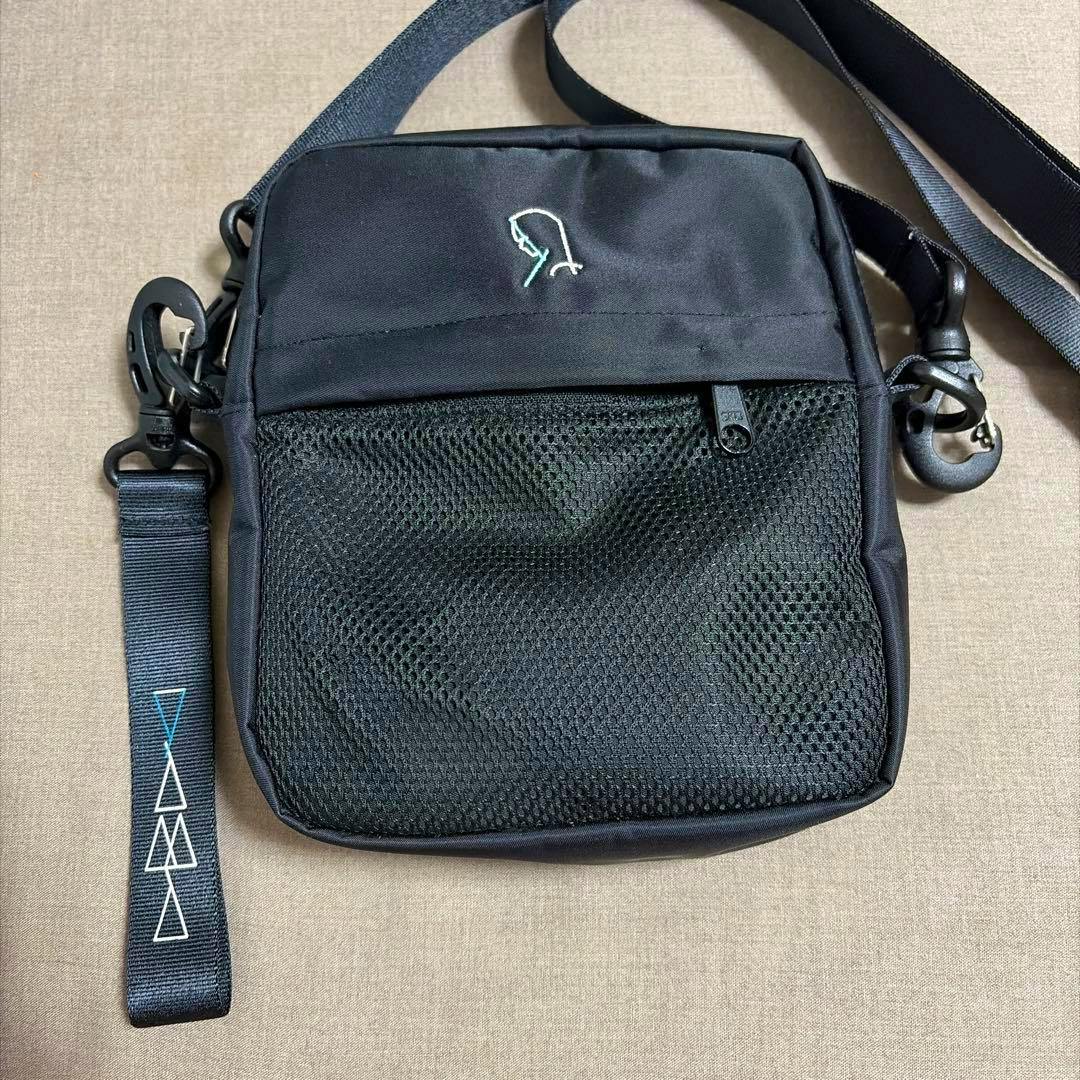 Front view of the yama アーティストグッズ ショルダーバッグ (black shoulder bag) showing a mesh front pocket, small embroidered logo, and attached strap with patterned tab.